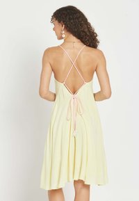 Mujer con cabello rizado que lleva un vestido sin mangas de color amarillo pálido con tirantes finos cruzados en rosa y un lazo anudado en la parte trasera.