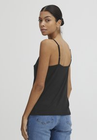 Zwarte cami-top met verstelbare bandjes, gemaakt van een gladde, lichte stof. In combinatie met een blauwe spijkerbroek staat het model met de rug naar de camera.