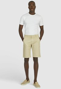 Beige katoenen shorts met een vlakke voorkant, twee zijzakken en een knoopsluiting, gecombineerd met een eenvoudige witte t-shirt met ronde hals en instap schoenen.