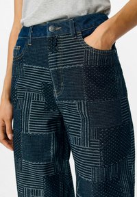 Pantalons en patchwork en denim bleu foncé avec des textures variées, comportant des rayures et des pois, des poches avant et une fermeture à bouton.