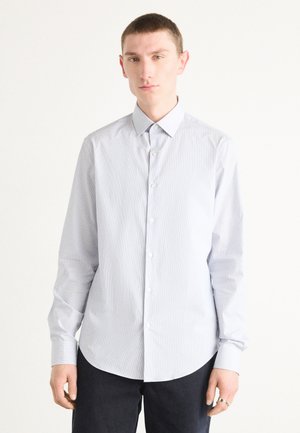 Camisa de manga larga color azul claro con un sutil patrón de rayas, cuello abotonado y dobladillo redondeado. Presenta botones lisos y una textura suave.