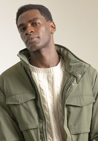 Chaqueta verde oliva con cuello alto, cierre de cremallera y bolsillos con solapa, superpuesta a un suéter de punto trenzado color crema. Se aprecian materiales texturizados.