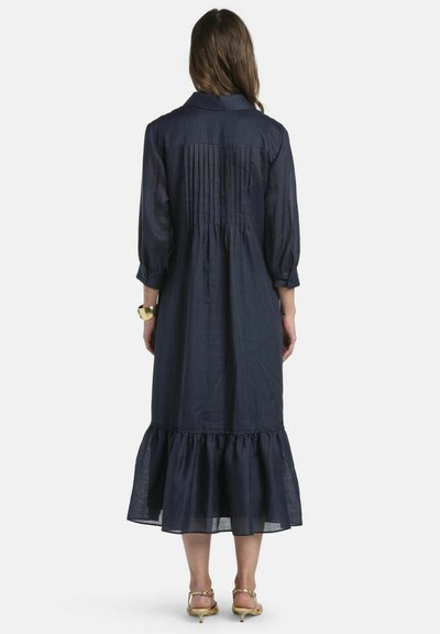 Žena v tmavomodrých midi šatách s plisovaným horným zadným dielom a volánovou lemovkou, doplnená zlatými pásikovými podpätkami a náramkom.
