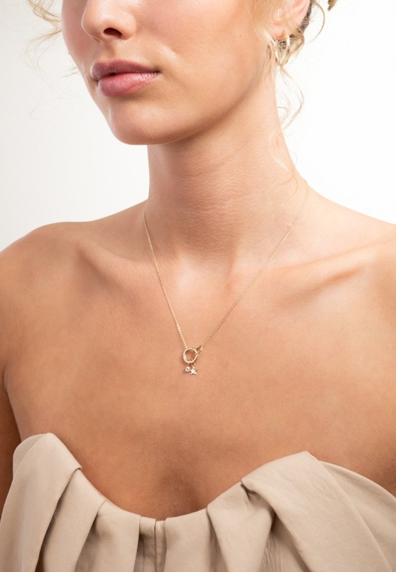Gouden ketting met een cirkelvormige hanger en een klein sterrenbedel, met een delicate ketting tegen een blote sleutelbeen en gestructureerde stof.