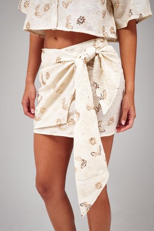 Vrouw draagt een crèmekleurige rok met bloemrijke borduursels en een grote gestrikte strik aan de zijkant, gecombineerd met een bijpassend cropped topje.