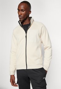 Felpa zip-up beige chiaro con colletto alto, dotata di una tasca sul petto e tessuto testurizzato. Abbinata a pantaloni cargo neri.