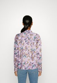 Donna con capelli scuri raccolti in una coda di cavallo, vestita con una camicia floreale viola a maniche lunghe e jeans blu, vista da dietro su uno sfondo bianco.