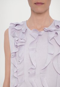 Gros plan d'une personne portant un haut sans manches violet clair avec un tissu texturé et des détails à volants superposés sur le devant.