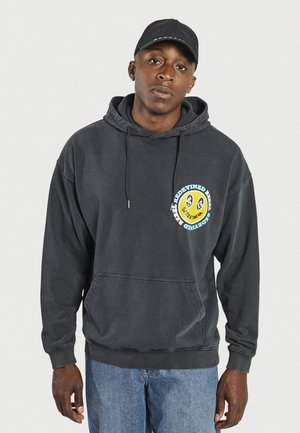 Sudadera negra con bolsillo canguro y detalles en gris, con un gráfico circular en azul y amarillo en el pecho, combinada con jeans azules.