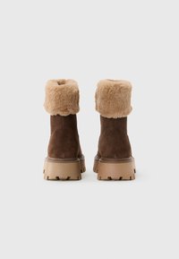 Botas de tobillo de ante marrón con gruesos puños de piel de imitación beige y suelas de goma gruesas de color marrón claro, vistas desde la parte posterior sobre un fondo blanco sencillo.