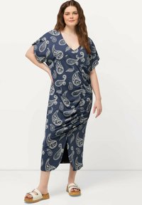 Laivansinis maxi-mekko, jossa on paisleymalli, V-pääntie, lyhyet hihat ja rypytetty yksityiskohta, yhdistettynä kömpelöihin valkoisiin sandaaleihin.