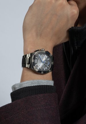 Pols met een horloge met een zwarte metalen band dat 10:08 aangeeft, gedragen over een grijze mouw en een donker bordeauxrode jas met een ritssluiting naast de pols.