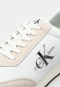 Fehér sneaker bézs velúr részletekkel, sima dizájnnal, kerek orral, fehér fűzőkkel és fekete logóval az oldalon.