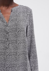 Vrouw in een zwart-wit gestreepte blouse met lange mouwen, versierd met kleine abstracte vormen en parelknoopjes bij de hals.