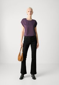Blusa viola a maniche corte, jeans neri a campana, stivaletti neri alla caviglia, che tiene una borsa a tracolla color beige. Tessuto morbido con maniche arrotolate.
