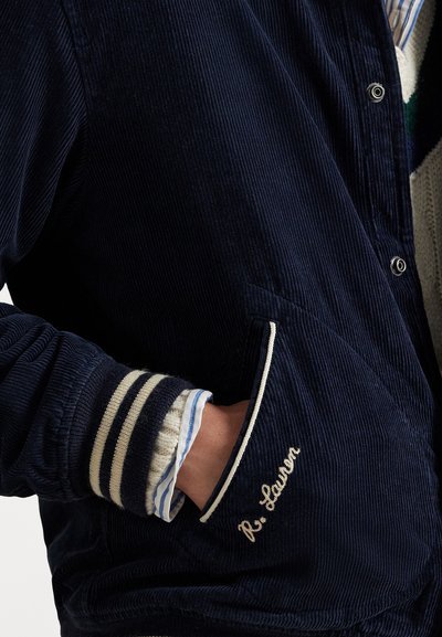 Hand in the pocket of a navy blue corduroy jacket with white striped ribbed cuffs and "R. Lauren" embroidered near the pocket.