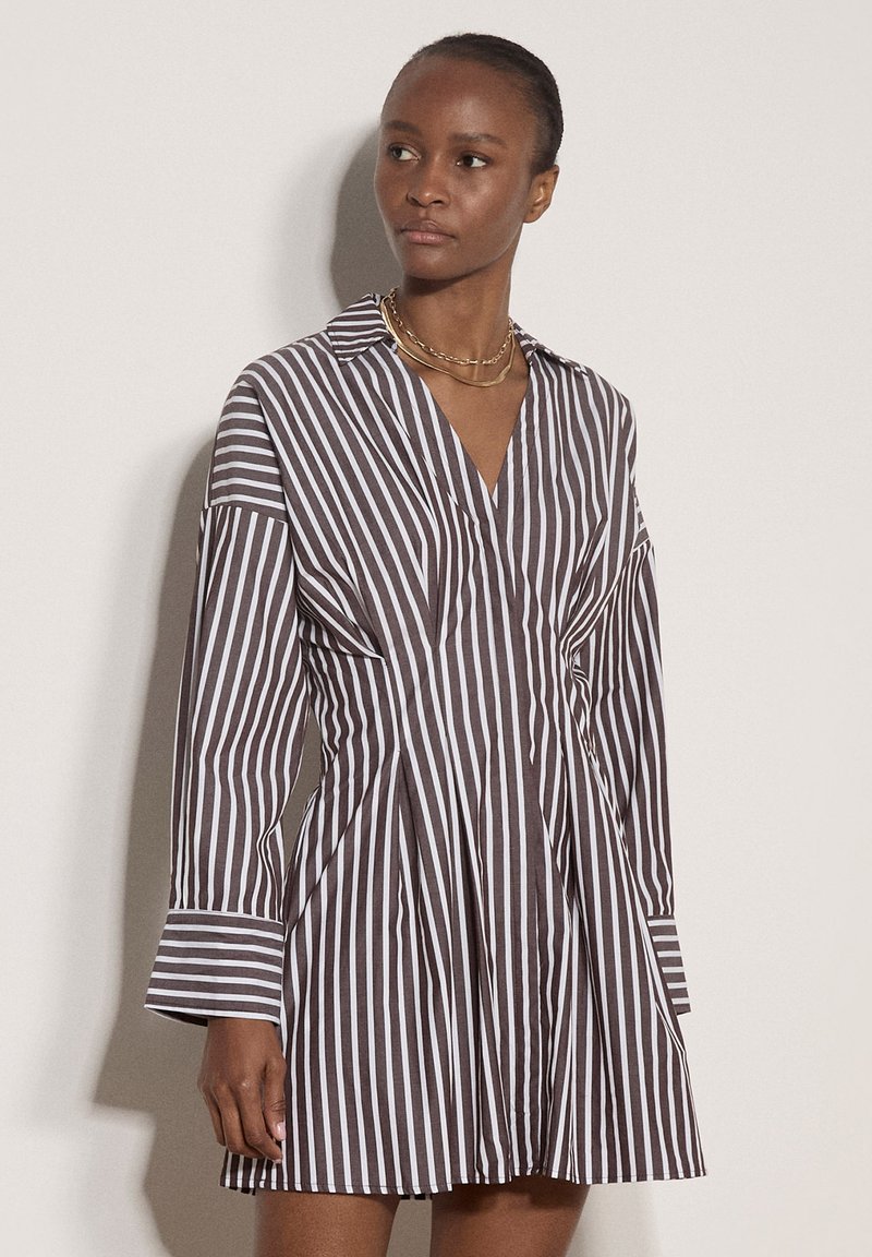 Femme portant une robe à rayures verticales marron et blanche avec manches longues et col, accessoirisée de colliers en or superposés.