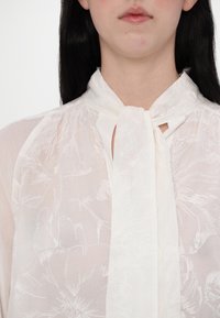 Blusa bianca con motivo floreale, morbida al tatto e con dettaglio al collo a lacci. Presenta un tessuto trasparente con una leggera lucentezza e cuciture delicate.