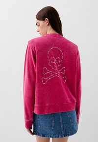 Felpa fucsia con un design di teschio e ossa incrociate in strass sul retro. Tessuto morbido, vestibilità rilassata, collettino e polsini a costine.