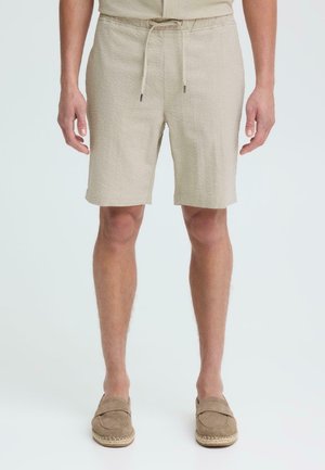 Mężczyzna w beżowych, teksturowanych szortach ze ściągaczem i pasujących mokasynach, stojący na tle białego tła.