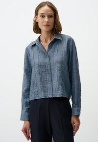 Lichtblauwe gestructureerde blouse met een cropped snit, knoopsluiting aan de voorkant en lange mouwen. Heeft een zachte, gekreukte stof en een kraag.