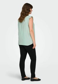 Lichtgroene, mouwloze top met gekante scalloped rand; gedragen met zwarte slim-fit jeans en zwarte flats, gezien vanaf de achterkant.