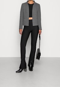 Donna con un blazer grigio, un top corto nero, pantaloni neri aderenti con aperture frontali, stivali neri con tacco, e che tiene una borsa nera con tracolla a catena.