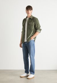 Olijfgroene corduroy shirt met twee borstzakken, wit T-shirt, blauwe jeans en witte sneakers met beige zolen. Eenvoudige, casual kleding.