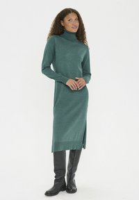 Femeie în picioare, îmbrăcată într-o rochie pulover midi cu guler înalt de culoare teal, cu crăpătură laterală și cizme negre până la genunchi, pe un fundal alb.