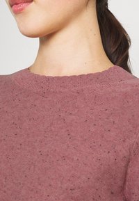 Pullover in staubigem Rosaton mit rundem Ausschnitt, strukturierter Strickoptik und dezenten schwarzen Sprenkeln. Aus weichem Stoff mit geripptem Kragen.