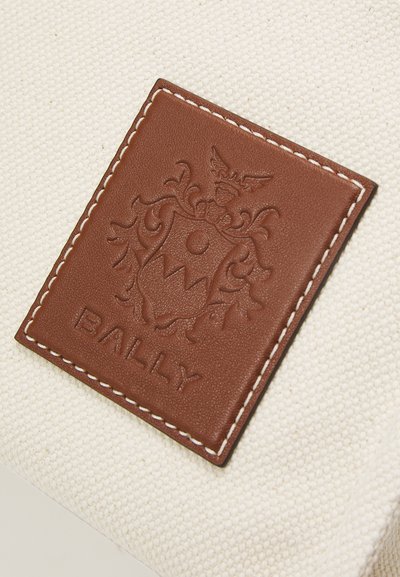 Barna bőr címke domború címerrel és fehér cérnával varrt "BALLY" felirattal, egy texturált, törtfehér szövet hátterére rögzítve.
