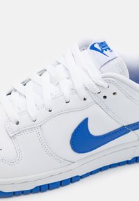 Valkoinen nahkainen Nike-tennari, jossa sininen Swoosh-logo, sininen pohja, valkoiset nauhat ja sininen Nike-brändäys kielekkeessä.