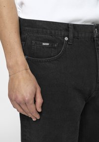 Zwarte denimjeans met een iets versleten textuur, met een klein logolabel op de voorkzak en een zilveren knoopsluiting. Hand ligt ernaast.