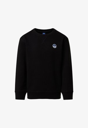 Jeune garçon les mains dans les poches, portant un sweat-shirt noir avec un logo, un short noir, des chaussettes blanches et des baskets blanches, debout sur un fond uni.