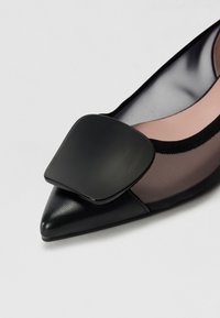 Ballerine noire à bout pointu avec un accent rectangulaire noir brillant et un revêtement en maille. Texture en cuir lisse avec un design léger.