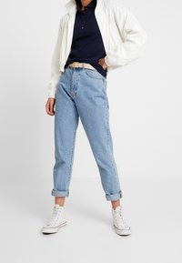 Veste zippée blanche, polo bleu marine, jean léger bleu clair retroussé et baskets montantes blanches. Look décontracté avec une ceinture mettant en valeur la taille.