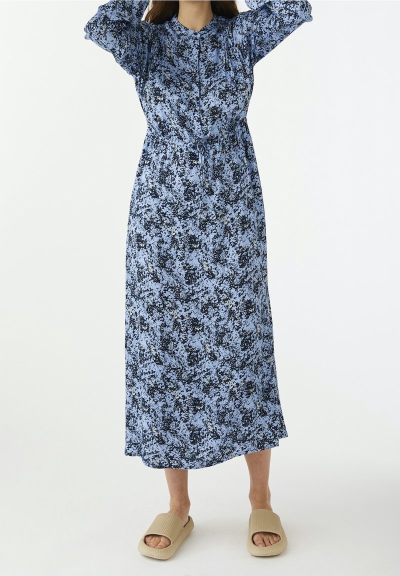 Blå blommig maxiklänning med långa ärmar, rynkad detalj och insvängd midja. Materialet verkar lätt och har en mjuk textur.