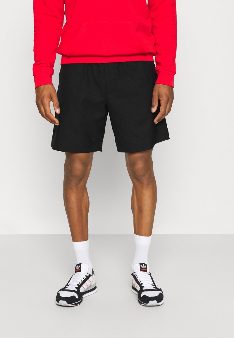 Homme portant un short noir, des chaussettes blanches, des baskets Adidas noires et blanches, et un sweat-shirt rouge, debout sur un sol clair et uni.