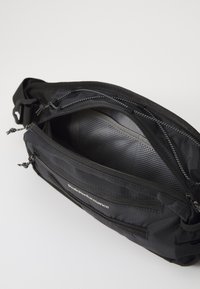 Čierna crossbody taška s viacerými zipsovými priehradkami. Má odolnú textíliu, štruktúrovanú podšívku a decentné značenie na prednej strane.