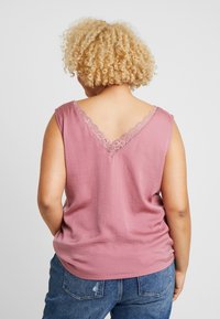Curly-haired person in a sleeveless pink top with lace trim at the back, wearing blue distressed denim jeans, standing against a plain background.