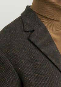 Blazer marrone intrecciato con una finitura testurizzata e rever a punta, abbinato a un maglione in maglia beige con collo alto.
