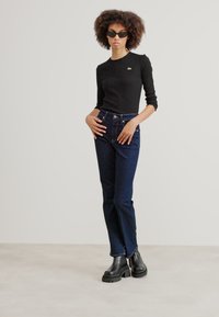 Svart langermet topp med en liten logo, mørkeblå høyhelsesjeans og kraftige svarte ankelstøvler. Enkel, tettsittende design, glatt tekstur.