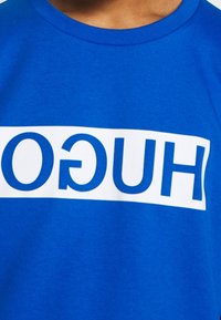 Modré bavlněné tričko s bílým horizontálním pruhem a textem "HUGO" tučnými modrými písmeny. Klasický kulatý výstřih a krátké rukávy.