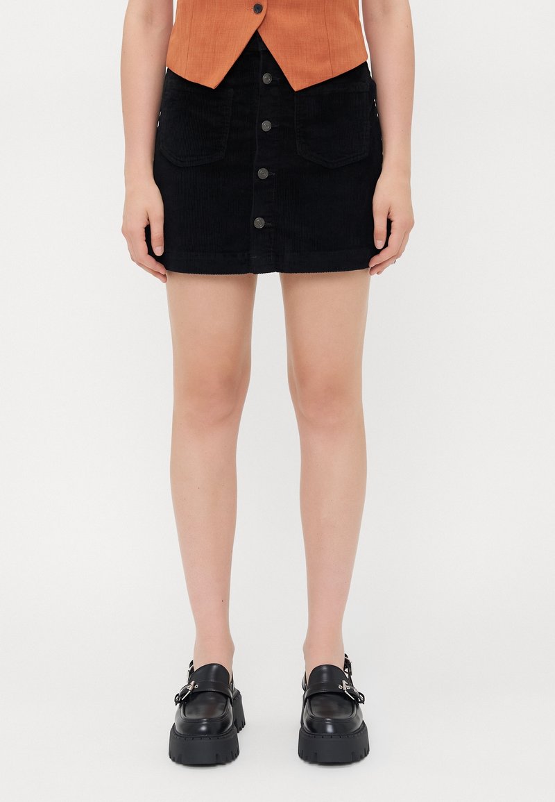 Femme portant une mini-jupe noire boutonnée avec des poches avant, des mocassins noirs épais et un top orange sans manches, debout sur fond blanc.