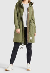 Personne portant une parka à capuche vert olive avec fermeture éclair et boutons-pression, chemise gris clair, pantalon noir et baskets blanches.