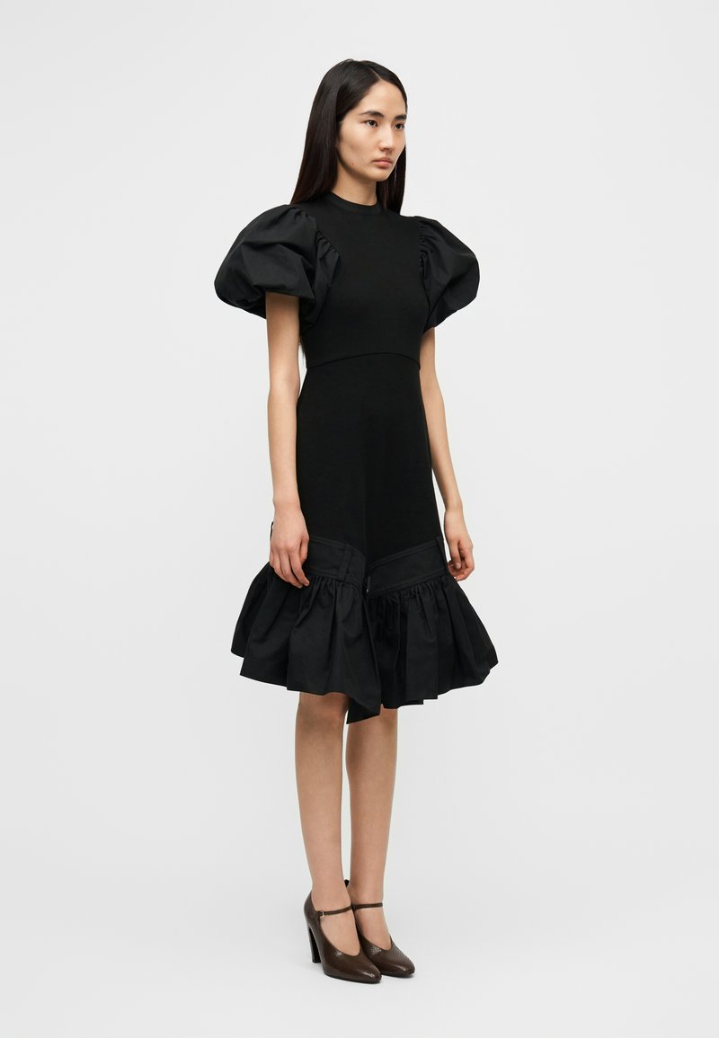 Vestito nero con maniche a sbuffo, corpetto aderente e orlo svasato con balza. Realizzato in tessuto strutturato con dettaglio a contrasto sull'orlo.