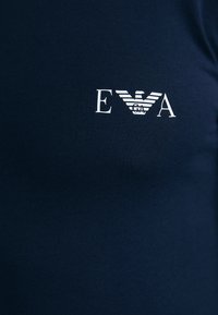 T-shirt in cotone blu navy con logo bianco contenente le lettere "EVA" e un design stilizzato di un'aquila sottostante. Texture liscia.