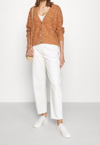 Žena v rezavě zbarveném zapínaném kardiganu, bílém topu, bílých kalhotách s vysokým pasem, bílých teniskách a s malou béžovou kabelkou přes rameno.