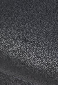 Zwart gestructureerd leer met een geperst "Calvin Klein" logo. Het oppervlak heeft een gepolijste textuur, wat zorgt voor een tactile ervaring.
