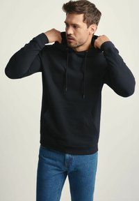 Mężczyzna poprawiający kaptur na czarnej bluzie z kapturem, ubrany w niebieskie dżinsy, na jednolitym tle.